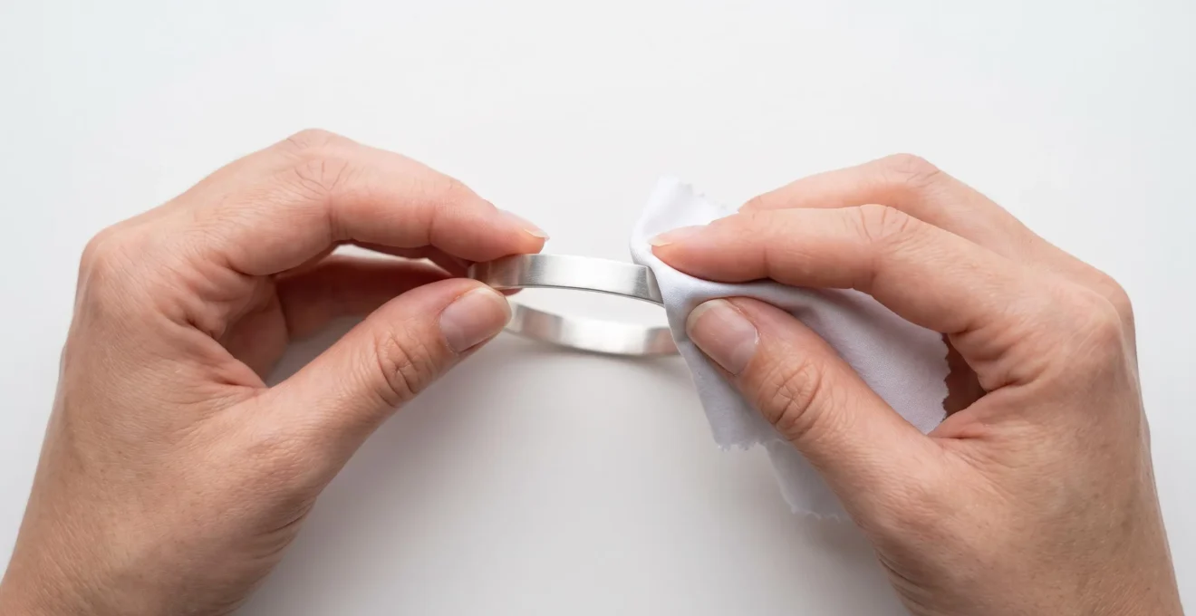 Gros plan sur deux mains nettoyant délicatement un bracelet jonc en argent avec un chiffon microfibre blanc sur surface blanche immaculée