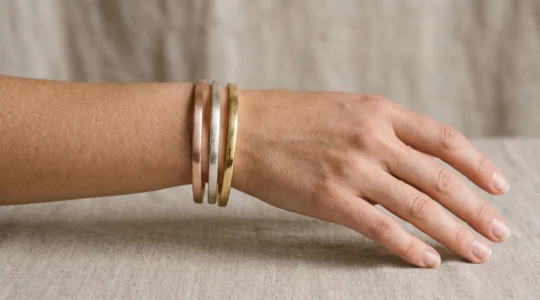 Gros plan sur un poignet portant trois bracelets joncs minimalistes superposés en or rose, argent brossé et or jaune sur fond beige neutre